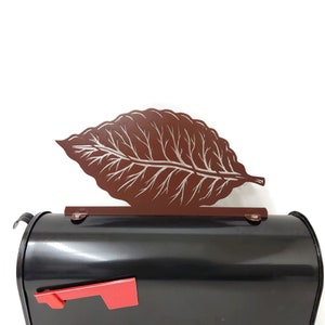 Peut inclure: Une boîte aux lettres noire avec un motif de feuille de tabac en métal brun sur le dessus. La boîte aux lettres a un drapeau rouge.