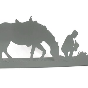 May include: A gray metal silhouette depicts a horse with a saddle, a kneeling figure, and a cross. The horse is grazing, and the figure is kneeling in prayer near the cross, suggesting a religious or memorial theme.