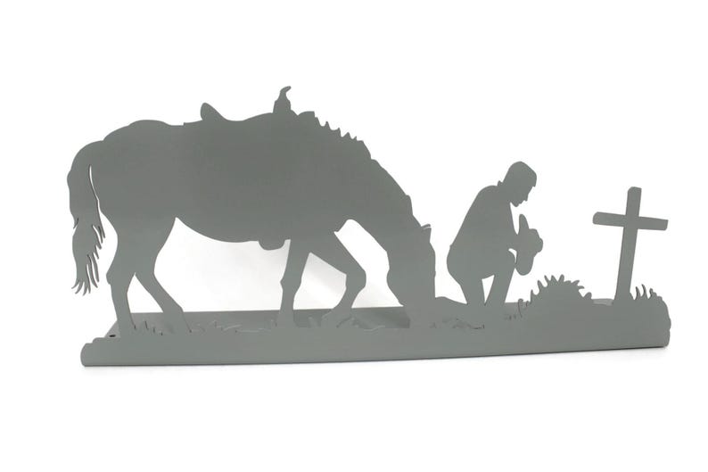 May include: A gray metal silhouette depicts a horse with a saddle, a kneeling figure, and a cross. The horse is grazing, and the figure is kneeling in prayer near the cross, suggesting a religious or memorial theme.