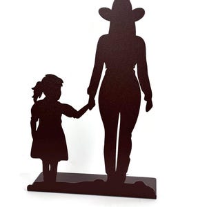 May include: A dark brown metal silhouette sculpture depicting a tall figure wearing a cowboy hat standing next to a smaller figure in a dress. The two figures are holding hands and are mounted on a flat, rectangular base of the same color.
