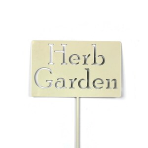 May include: A rectangular, cream-colored garden marker with the words "Herb Garden" cut out. The sign is mounted on a thin, pale-yellow stake, suitable for identifying herbs in a garden. The background is plain white.