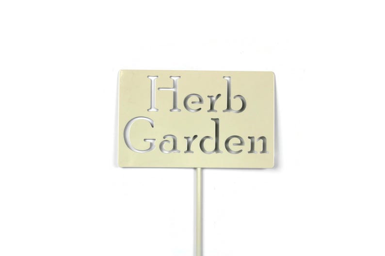 May include: A rectangular, cream-colored garden marker with the words "Herb Garden" cut out. The sign is mounted on a thin, pale-yellow stake, suitable for identifying herbs in a garden. The background is plain white.