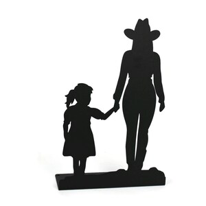 May include: A black metal silhouette sculpture depicting a tall figure wearing a cowboy hat standing next to a smaller child. The two figures are holding hands, and the entire piece is mounted on a flat, rectangular black metal base.