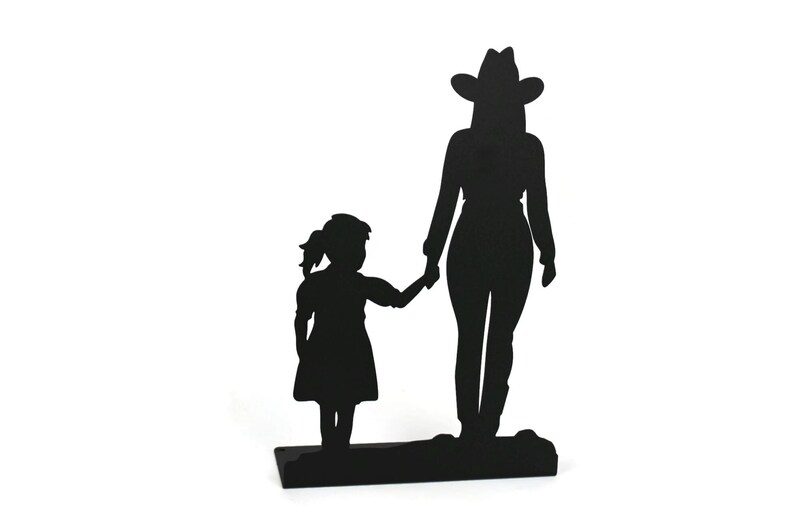 May include: A black metal silhouette sculpture depicting a tall figure wearing a cowboy hat standing next to a smaller child. The two figures are holding hands, and the entire piece is mounted on a flat, rectangular black metal base.