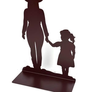May include: A dark brown metal silhouette sculpture featuring a tall figure wearing a cowboy hat holding the hand of a smaller child. The figures stand on a flat rectangular base, creating a decorative piece with a solid, uniform color finish.