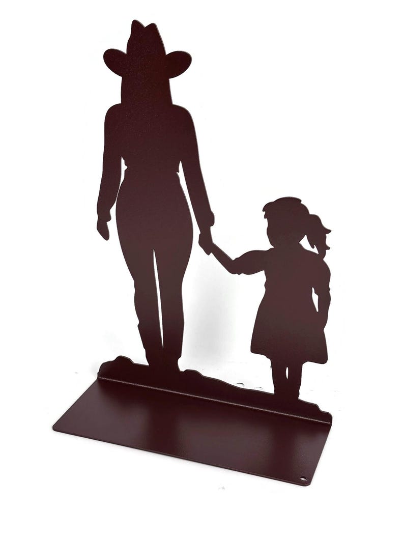 May include: A dark brown metal silhouette sculpture featuring a tall figure wearing a cowboy hat holding the hand of a smaller child. The figures stand on a flat rectangular base, creating a decorative piece with a solid, uniform color finish.