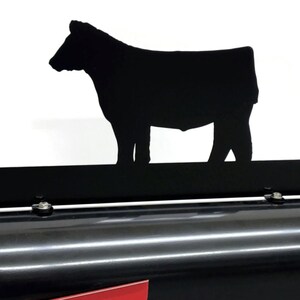 May include: Black metal cow silhouette mailbox topper. The topper features a detailed cutout of a cow standing on a rectangular bar. The mailbox is black with a red base.