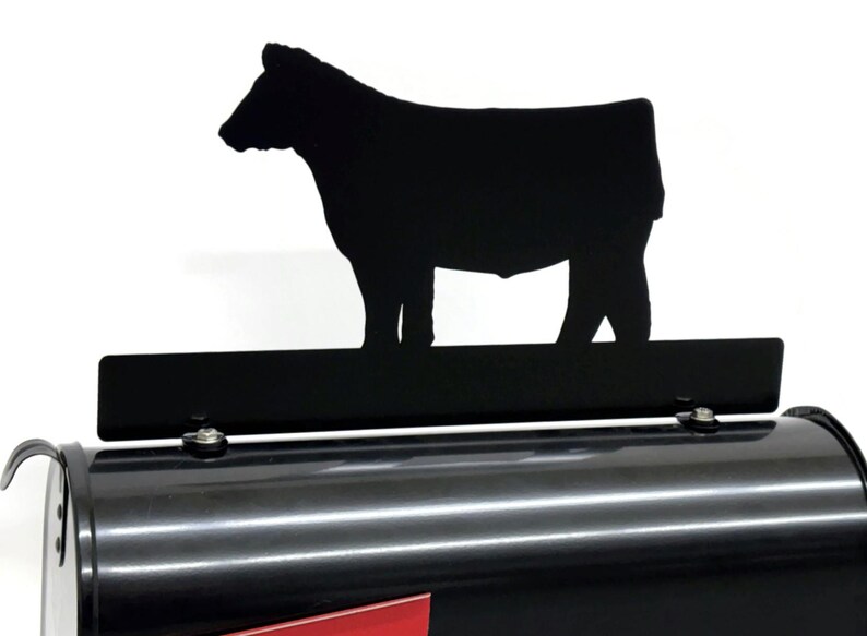 May include: Black metal cow silhouette mailbox topper. The topper features a detailed cutout of a cow standing on a rectangular bar. The mailbox is black with a red base.