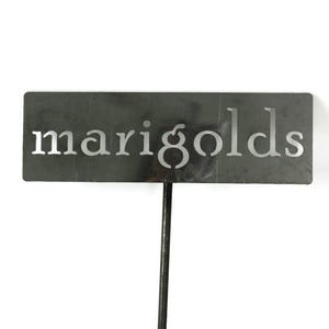 May include: A rectangular, dark gray metal garden stake with the word "marigolds" cut out in a sans-serif font. The stake is on a white background, ideal for marking flower beds.