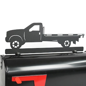 May include: A black metal pickup truck silhouette mounted on a black mailbox. The truck features a flatbed design and is attached to the mailbox with visible hardware. The mailbox has a red flag and a curved handle.