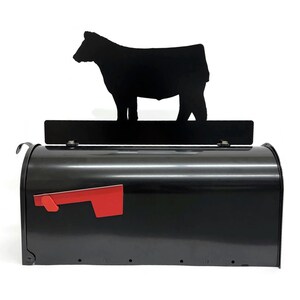 May include: A black metal mailbox with a red flag, topped with a black cow silhouette. The mailbox has a curved lid and a rectangular base. The cow is a simple, cut-out design, adding a rural touch to the mailbox.