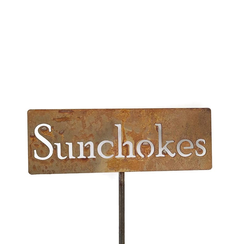 May include: A rectangular metal garden stake with the word "Sunchokes" cut out in a white font. The stake has a rusty brown finish and is mounted on a thin metal rod.