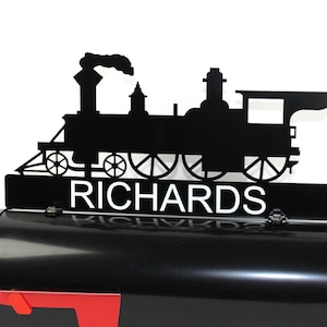 May include: Black metal train-shaped mailbox topper with the name "RICHARDS" in white lettering. The topper is mounted on a black mailbox with a red flag. The train silhouette includes details like wheels and a smokestack.