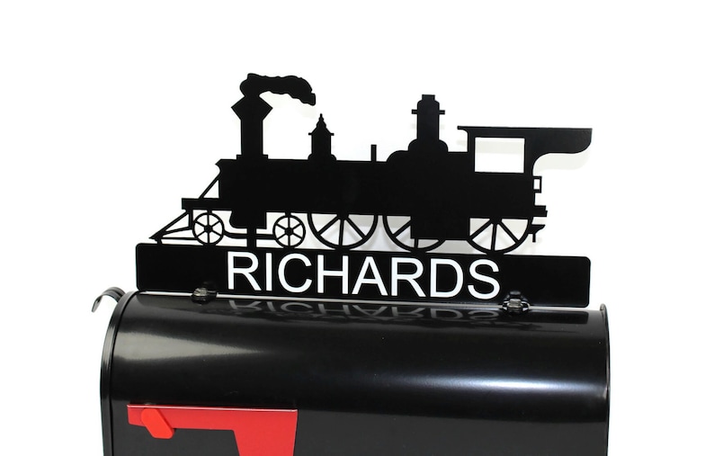 May include: Black metal train-shaped mailbox topper with the name "RICHARDS" in white lettering. The topper is mounted on a black mailbox with a red flag. The train silhouette includes details like wheels and a smokestack.