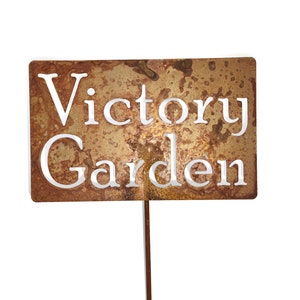 May include: A rectangular, weathered metal garden stake with the words "Victory Garden" cut out in white. The stake has a brown, rust-like appearance and is designed to be placed in a garden.