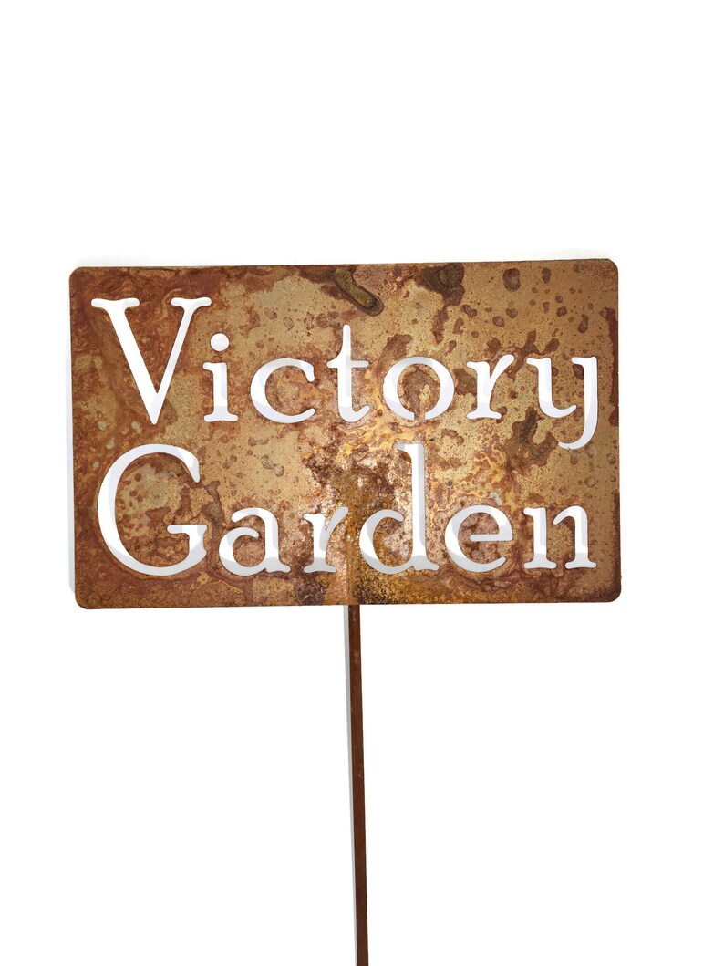 May include: A rectangular, weathered metal garden stake with the words "Victory Garden" cut out in white. The stake has a brown, rust-like appearance and is designed to be placed in a garden.