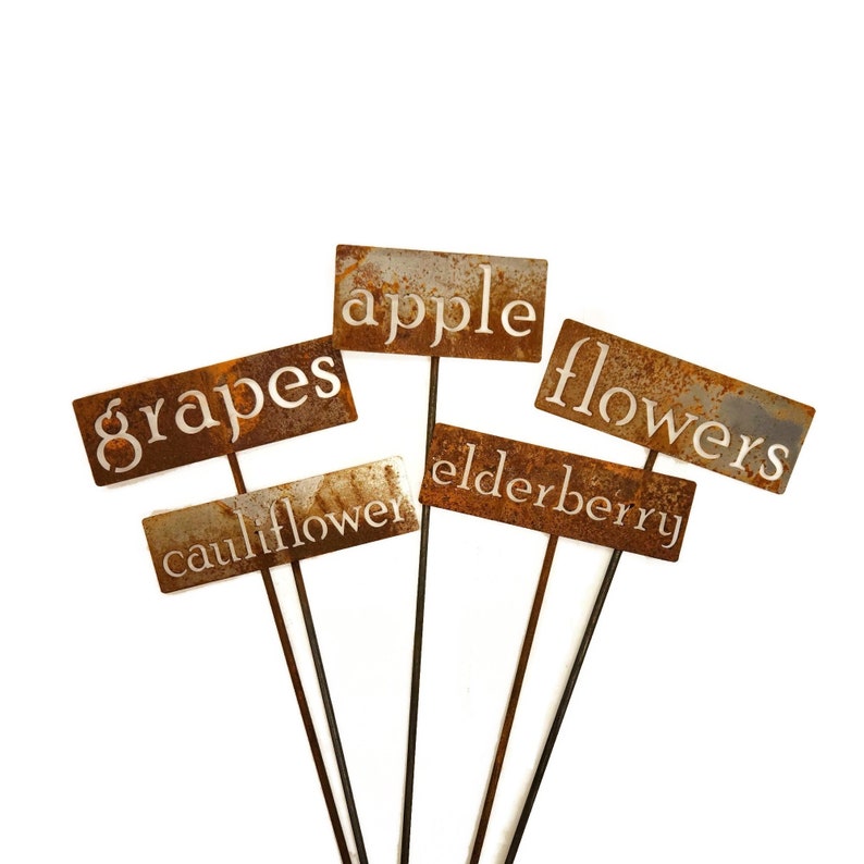 May include: A collection of rustic metal garden markers with text cutouts. The markers are rectangular with a weathered, rusty finish. The text includes "grapes", "apple", "flowers", "cauliflower", and "elderberry".