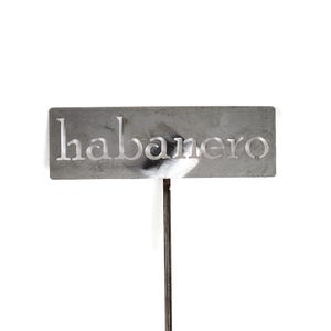 May include: A rectangular, gray metal garden marker with the word "habanero" cut out in white letters. The marker is attached to a thin, dark metal stake, suitable for labeling plants in a garden.