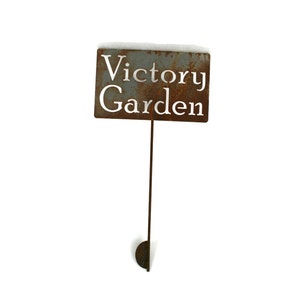 May include: A rectangular, weathered metal garden sign with the words "Victory Garden" cut out. The sign is mounted on a thin, rusty metal stake with a half-circle base. The top of the sign has a dusting of snow.