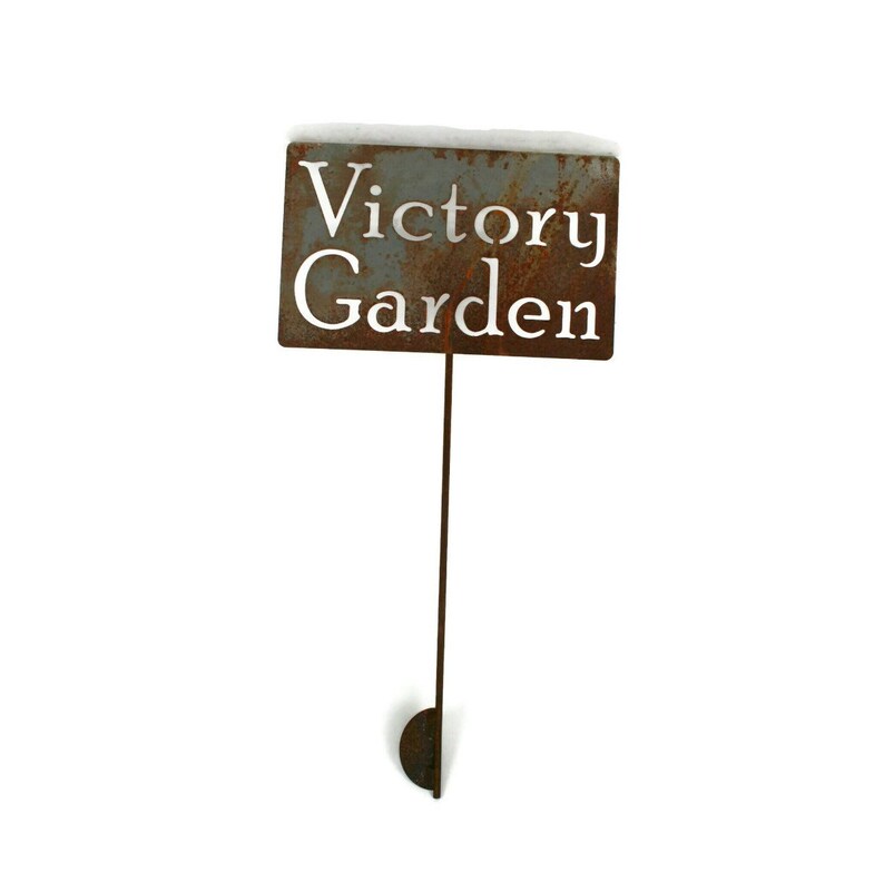 May include: A rectangular, weathered metal garden sign with the words "Victory Garden" cut out. The sign is mounted on a thin, rusty metal stake with a half-circle base. The top of the sign has a dusting of snow.