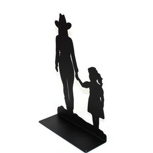 May include: A black metal silhouette sculpture featuring an adult wearing a cowboy hat standing next to a child. The two figures are holding hands, and the entire piece is mounted on a flat, rectangular black metal base for stability.