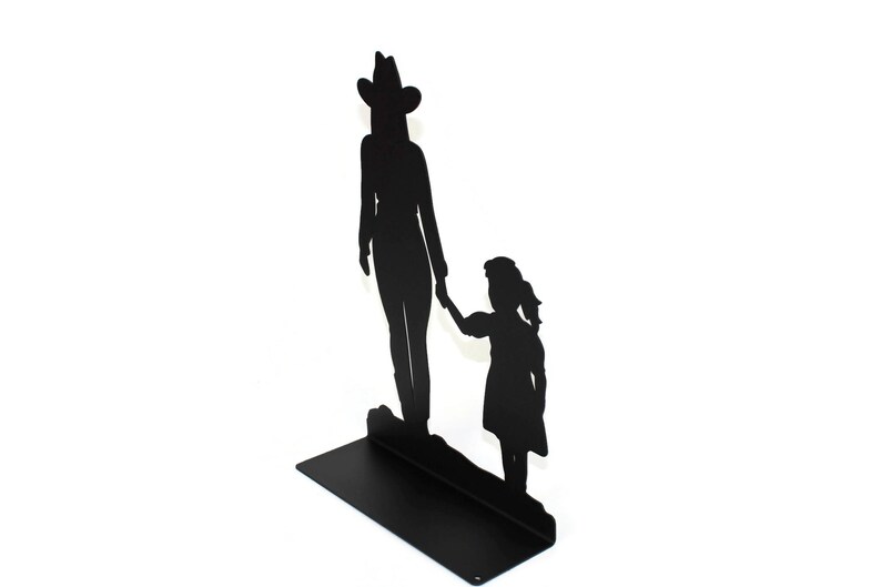 May include: A black metal silhouette sculpture featuring an adult wearing a cowboy hat standing next to a child. The two figures are holding hands, and the entire piece is mounted on a flat, rectangular black metal base for stability.