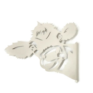 May include: A white metal wall decor in the shape of a cow's head. The cow's head is facing the right side of the image and has a playful expression. The cow's head is cut out of the metal, creating a silhouette effect.