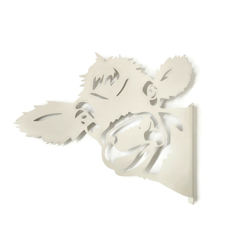 May include: A white metal wall decor in the shape of a cow's head. The cow's head is facing the right side of the image and has a playful expression. The cow's head is cut out of the metal, creating a silhouette effect.