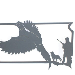 May include: A dark gray metal wall art piece depicting a pheasant in flight, a hunter with a rifle, and a dog. The artwork is cut from metal and mounted on a white background. The pheasant is in mid-flight, with its wings spread wide.