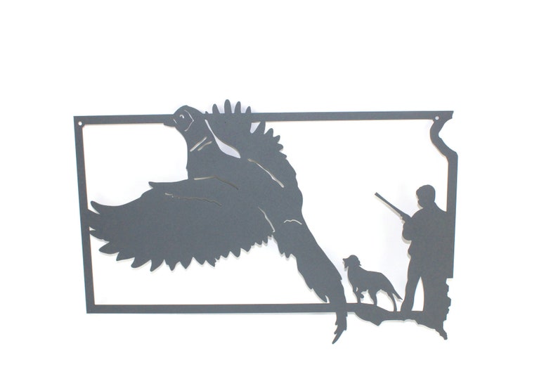 May include: A dark gray metal wall art piece depicting a pheasant in flight, a hunter with a rifle, and a dog. The artwork is cut from metal and mounted on a white background. The pheasant is in mid-flight, with its wings spread wide.