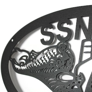 Puede incluir: Un letrero de pared de metal negro mate con un marco ovalado y el texto SSN 7, acompa&ntilde;ado de una ilustraci&oacute;n recortada detallada de una criatura marina con patrones intrincados y una forma de ancla estilizada integrada en el dise&ntilde;o.