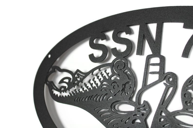 Puede incluir: Un letrero de pared de metal negro mate con un marco ovalado y el texto SSN 7, acompa&ntilde;ado de una ilustraci&oacute;n recortada detallada de una criatura marina con patrones intrincados y una forma de ancla estilizada integrada en el dise&ntilde;o.