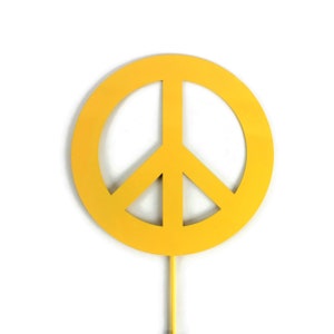 May include: A bright yellow peace sign on a stick, set against a white background. The peace symbol is a circle with three lines inside, representing peace and harmony. The item is likely a decorative piece or a cake topper.