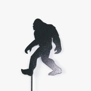 May include: A black and silver glitter Bigfoot silhouette on a thin black stick. The figure is walking, with one leg forward. The background is plain white.