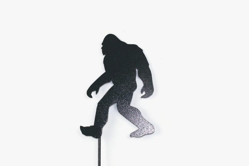 May include: A black and silver glitter Bigfoot silhouette on a thin black stick. The figure is walking, with one leg forward. The background is plain white.