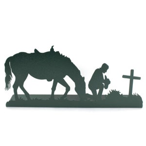 May include: A dark green metal silhouette depicts a horse, a kneeling figure, and a cross. The horse is grazing, the figure is kneeling with hands clasped, and the cross stands nearby. The scene evokes a sense of faith and reverence.