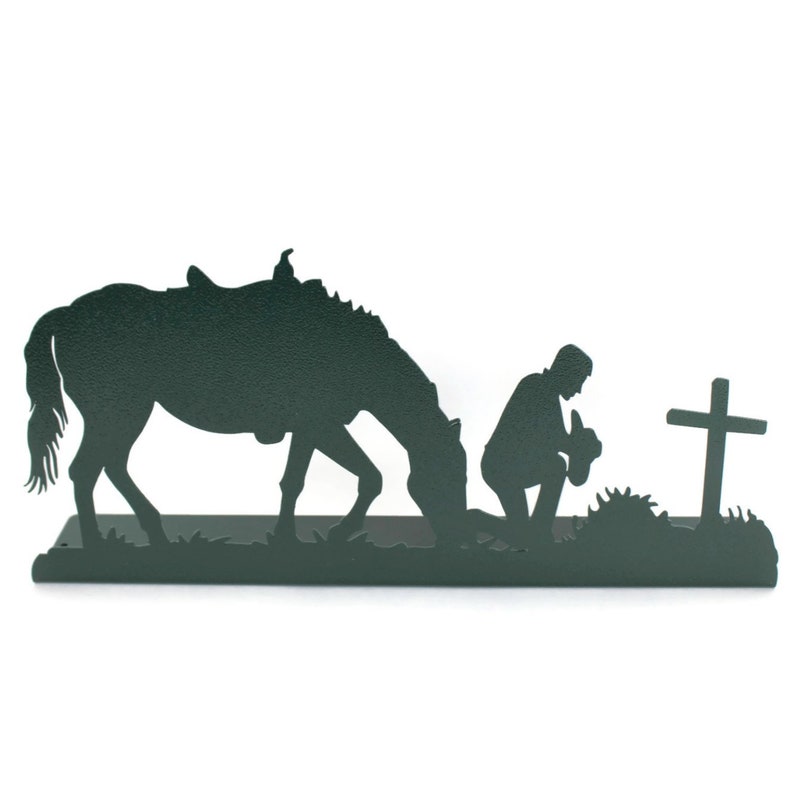 May include: A dark green metal silhouette depicts a horse, a kneeling figure, and a cross. The horse is grazing, the figure is kneeling with hands clasped, and the cross stands nearby. The scene evokes a sense of faith and reverence.
