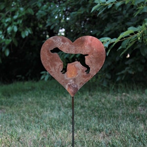May include: A heart-shaped, rusty metal garden stake featuring a silhouette of a dog. The dog is positioned within the heart's center. The stake is placed in a grassy area with a backdrop of green foliage.