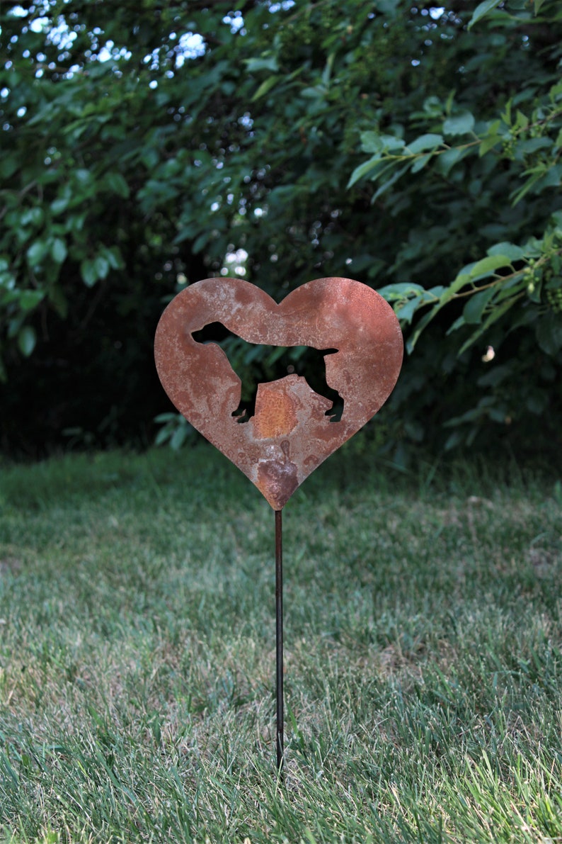 May include: A heart-shaped, rusty metal garden stake featuring a silhouette of a dog. The dog is positioned within the heart's center. The stake is placed in a grassy area with a backdrop of green foliage.