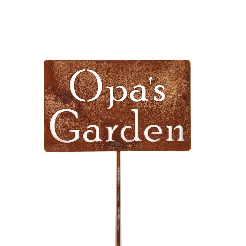 May include: A rectangular, rusty brown metal garden sign with the words "Opa's Garden" cut out in white lettering. The sign is mounted on a thin, metal stake, perfect for a garden or yard. The sign is designed to be placed in the ground.