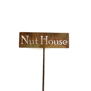 May include: A rectangular metal sign with the words "Nut House" cut out. The sign has a rusty brown finish and is mounted on a thin, dark metal stake, suitable for garden or yard decor.