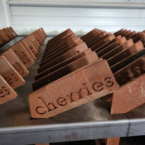 May include: A close-up of a stack of rusty metal signs with the word "cherries" etched into one of the signs. The signs are arranged in a staggered pattern, creating a visual texture.