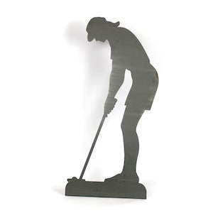 May include: A dark gray metal silhouette of a person playing golf. The figure is bent over, holding a golf club, and wearing a hat and shorts. The silhouette is mounted on a rectangular base.