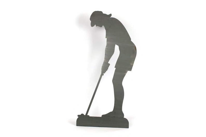 May include: A dark gray metal silhouette of a person playing golf. The figure is bent over, holding a golf club, and wearing a hat and shorts. The silhouette is mounted on a rectangular base.