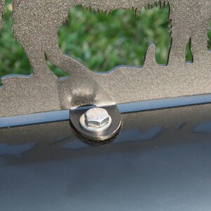 Puede incluir: Primer plano de un buz&oacute;n de metal con una tapa decorativa de metal que presenta la silueta de tres animales. El buz&oacute;n es negro y la tapa es plateada. Se ve un tornillo plateado en el buz&oacute;n.