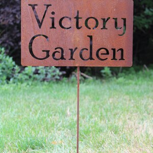 May include: A rectangular, rusty metal garden sign with the words "Victory Garden" cut out. The sign is mounted on a thin, rusty metal stake, and is placed in a grassy area.