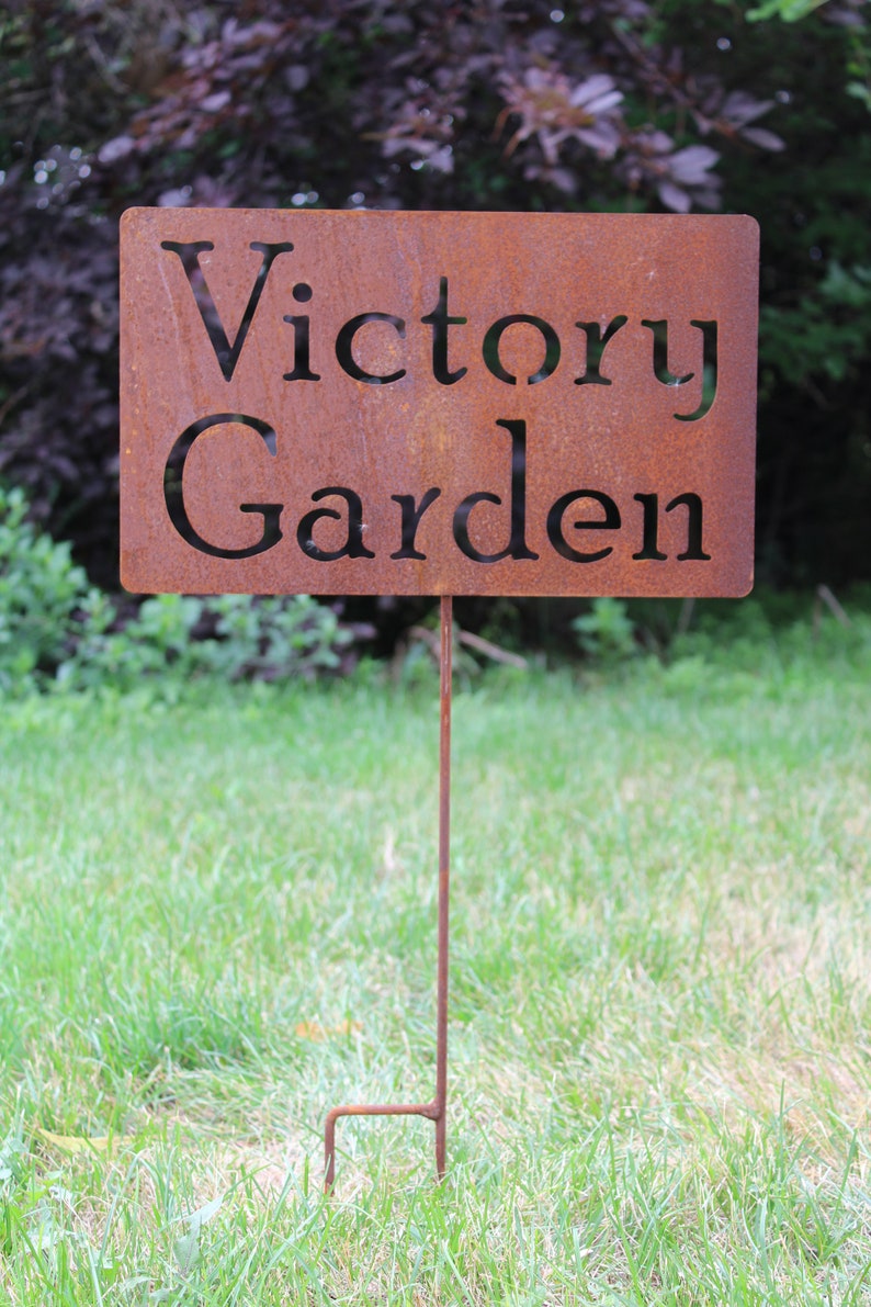 May include: A rectangular, rusty metal garden sign with the words "Victory Garden" cut out. The sign is mounted on a thin, rusty metal stake, and is placed in a grassy area.