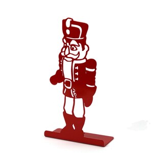 May include: Red metal nutcracker figurine with a white face and black details. The nutcracker is standing on a red metal base.