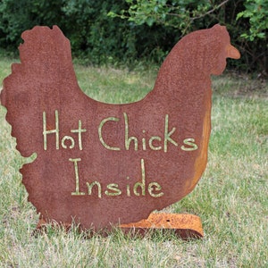 May include: A rusty metal chicken silhouette with the words "Hot Chicks Inside" written on it. The chicken is standing on a grassy lawn.
