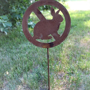 May include: A rusty metal sign with a circle and a diagonal line through it. The sign has a silhouette of a rabbit inside the circle. The sign is on a metal stake and is stuck in the ground.